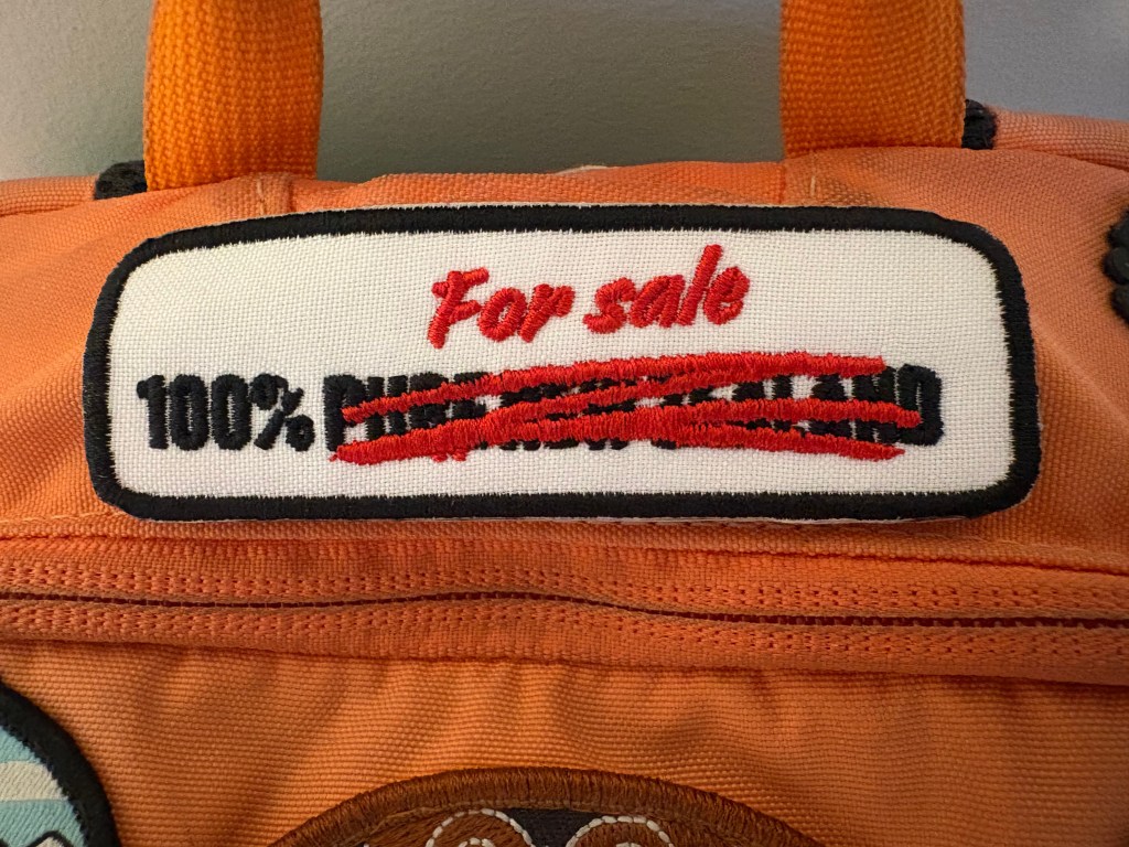 A close-up of an orange bag featuring a patch that reads 'For sale' with '100% PURE' crossed out, indicating a humorous or ironic message.